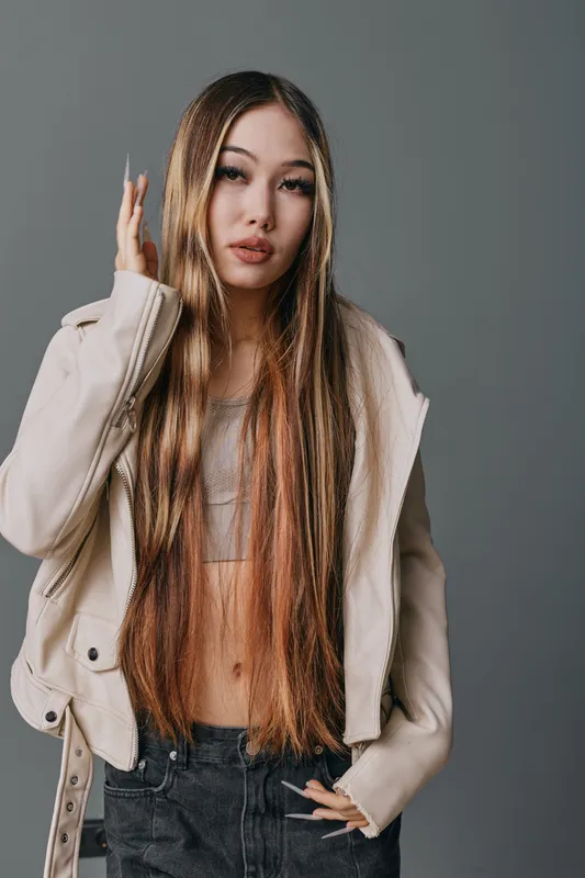 Young woman with long straight hair and casual outfit posing against gray background. Fashion model with natural makeup in beige jacket and black pants for portrait photography.