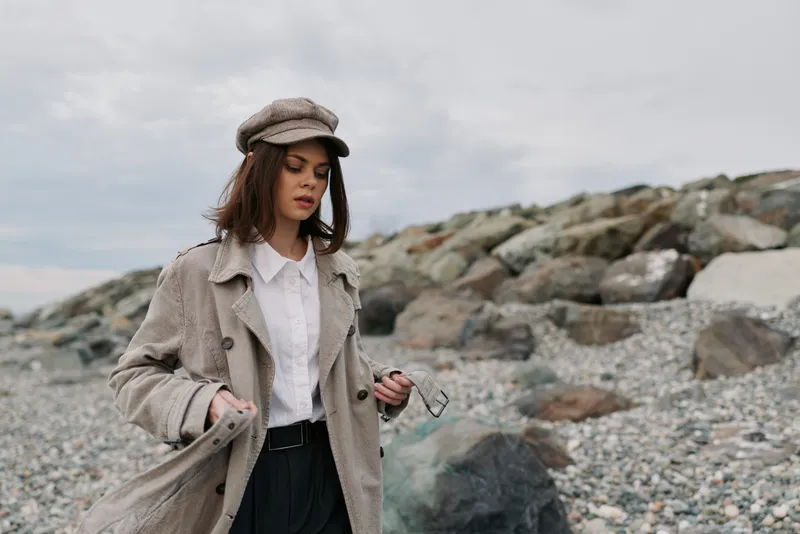 Young woman in beige coat and hat walks on rocky beach under cloudy sky. Casual autumn fashion with natural makeup and calm mood in outdoor coastal landscape scene.