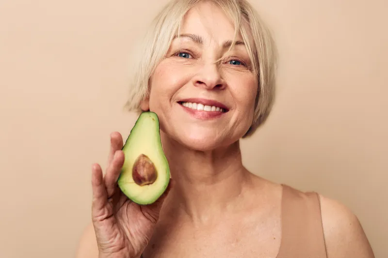 Smiling mature woman with short blonde hair holds fresh avocado half close to face on beige background. Concept of healthy lifestyle, natural food, nutrition, and wellness for older adults.