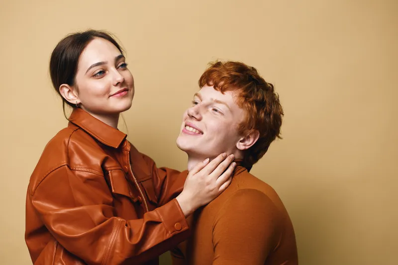 Couple with affectionate body language posing in warm brown clothing on beige background. Young man and woman showing love and gentle connection with smiles in studio.