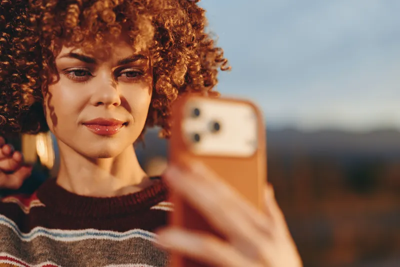 Woman selfie smartphone curly hair outdoors portrait sunset lifestyle, holding a phone for a casual self portrait, warm evening light, knit sweater, soft smile, blurred hills background