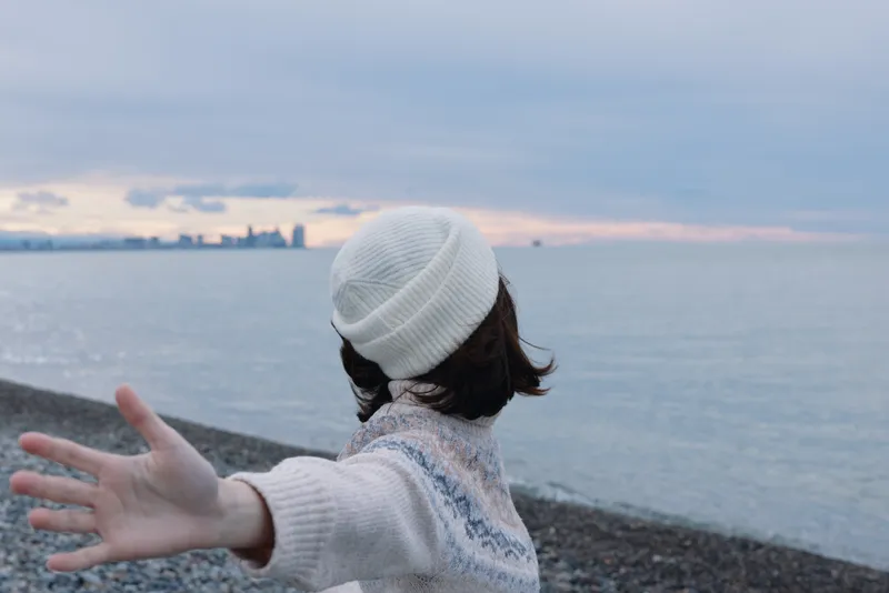 Woman standing on a pebble beach with outstretched arms, wearing a white beanie and knitted sweater, facing a calm sea as the sunset paints the horizon in soft colors.