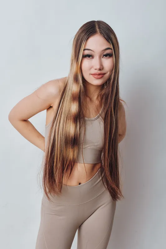 Young woman with long straight hair wearing beige sportswear stands against white background, smiling and posing with hands on hips for portrait photo.