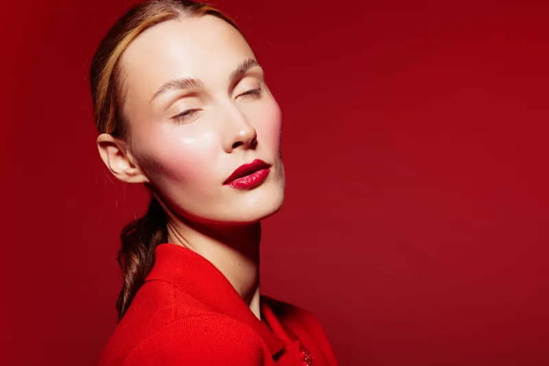 Portrait of a young woman with closed eyes wearing red lipstick and a red outfit against a red background. Concept of beauty, fashion, makeup, and elegance.