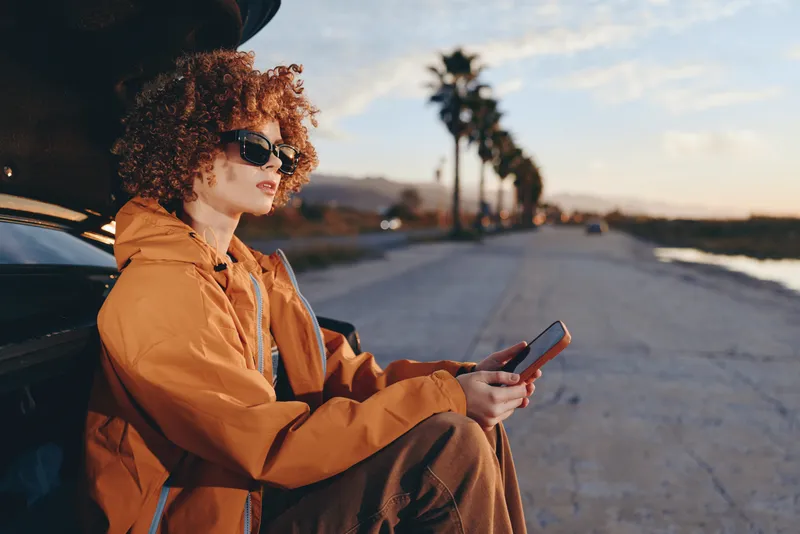 Woman lifestyle, curly hair, sunglasses, orange jacket, sitting near car on road, holding smartphone, relaxed travel mood, warm sunset light, outdoor street background