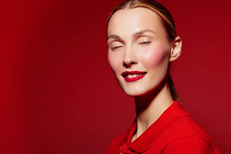Close-up of a smiling woman with red lipstick and red outfit posing with eyes closed on red background. Concept of beauty, fashion, makeup, and confident female model portrait.