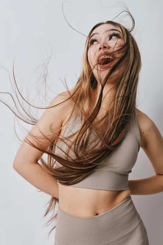 Young woman with long hair wearing a crop top and skirt smiles joyfully with hair flowing in the air against plain background. Fashion, beauty and happiness concept.