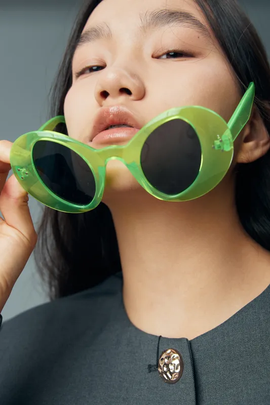 Woman with clear skin holding green round sunglasses near lips, wearing dark clothing with metallic button, closeup portrait with straight black hair in minimalistic studio light.