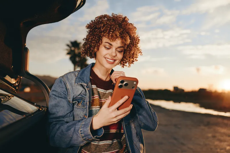 Woman lifestyle smartphone car sunset outdoor travel smiling at golden hour as she stands by an open car door holding a phone and looking at the screen warm casual denim jacket concept modern living