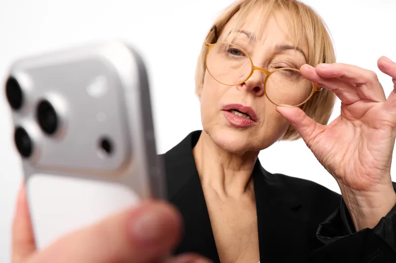Senior woman with glasses adjusting frames near smartphone, technology and communication selfie on white background, portrait, surprised expression focused