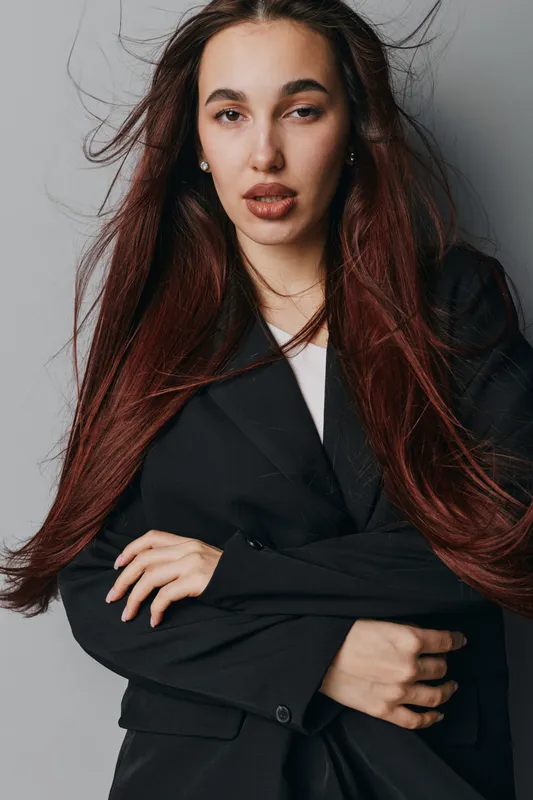 Woman with long hair and serious expression wearing black blazer posing with crossed arms on gray background. Fashion portrait, confident look, studio lighting, casual style and elegance.