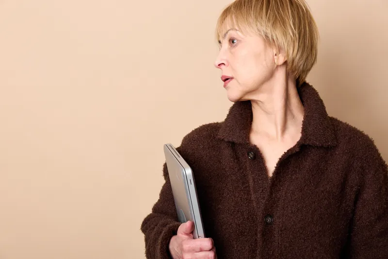Elderly woman holds smartphone in indoor sideview portrait, wearing fur coat, looking with concern about communication and technology, Neutral background