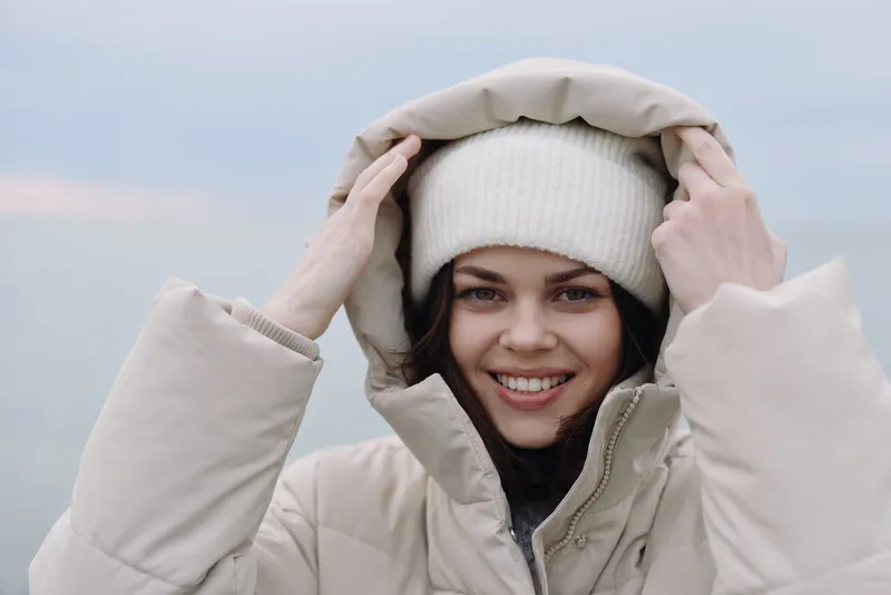 Winter scene with a smiling woman wearing a beige coat and cozy beanie, adjusting her hood outdoors. Clear sky and soft light highlight warmth, comfort, and winter fashion