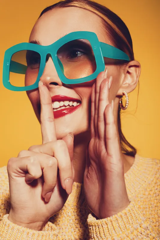 Young woman with large turquoise sunglasses and red lipstick smiles while making a shushing gesture on yellow background. Concept of fashion, style, and playful mood.