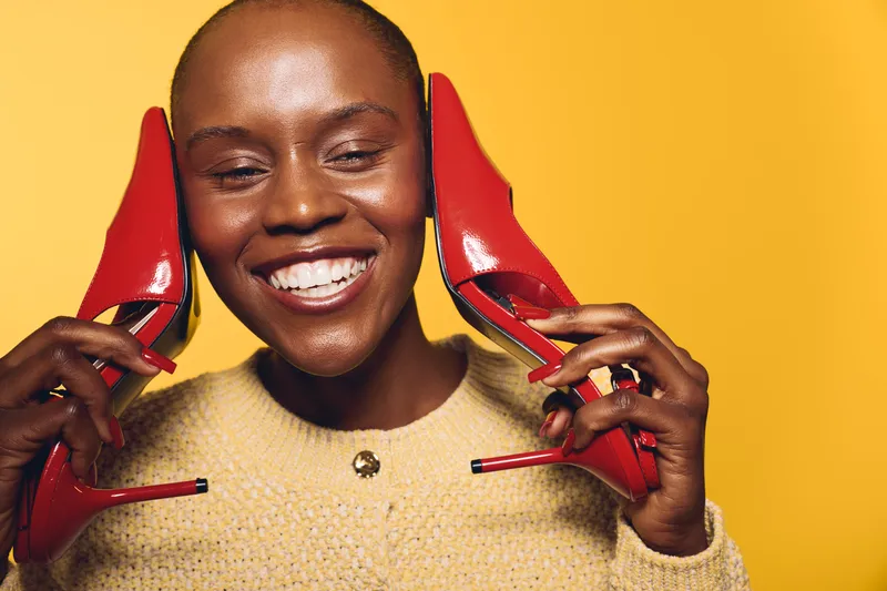 Smiling black woman with dark skin holds red high heels near face on yellow background. Concept of fashion, beauty, joy, and confident stylish female model with vibrant colors and accessories.