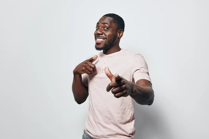 African American man with dark skin smiling and pointing fingers in casual light pink t shirt on white background. Positive gesture and confident expression.