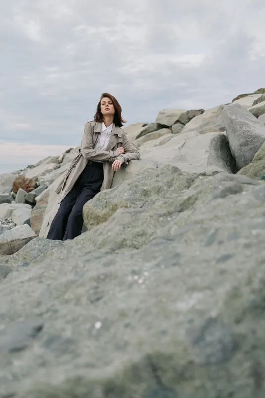Young woman in trench coat and black pants leans on large rocks under cloudy sky. Outdoor fashion portrait with natural stone background and relaxed mood by the sea.