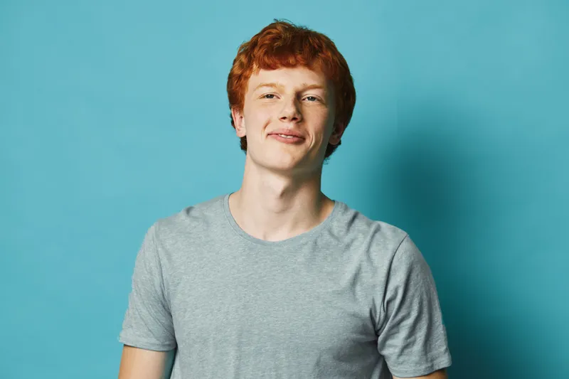 Young man with red hair and gray t shirt smiling and looking at camera on blue background in studio portrait with casual style and positive expression.