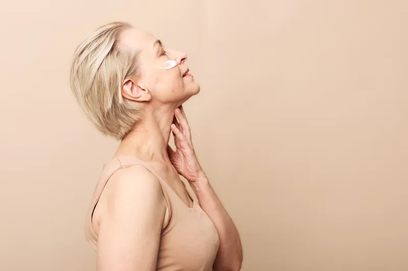 Mature woman with short blonde hair applies cream on face, eyes closed and relaxed, beige background. Concept of skincare, self-care, natural beauty, and aging gracefully.