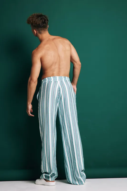 Young man with bare back wearing striped loose pants and white sneakers standing against green background in studio. Casual fashion, summer style and relaxed pose for male model.