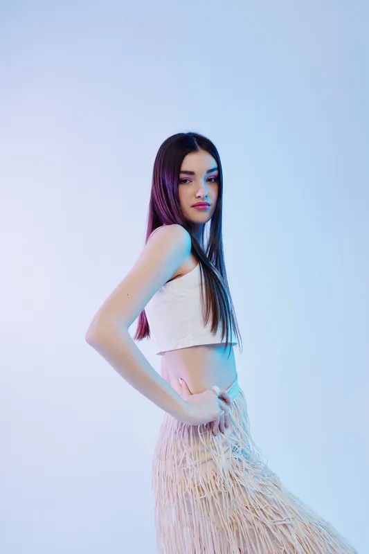 Young woman with long hair poses confidently in a white crop top and fringed skirt. Studio shot with soft blue lighting. Concept of woman beauty fashion and style.