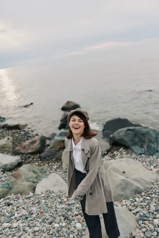 Happy young woman in casual outfit stands on rocky shore near calm sea, smiling and enjoying fresh air. Concept of outdoor lifestyle, nature, freedom, and positive mood by the water.