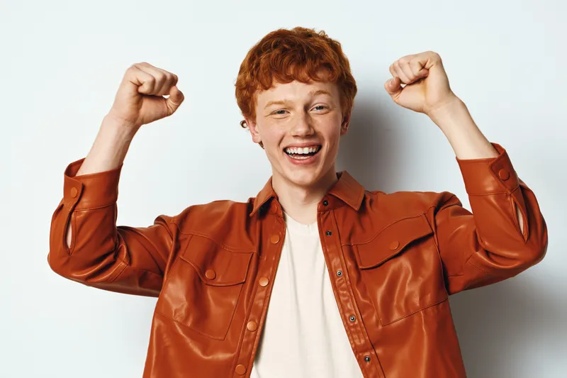Young man with red hair wearing brown leather jacket raising fists in excitement smiling on white background. Male, happy, gesture, victory, success, emotion.