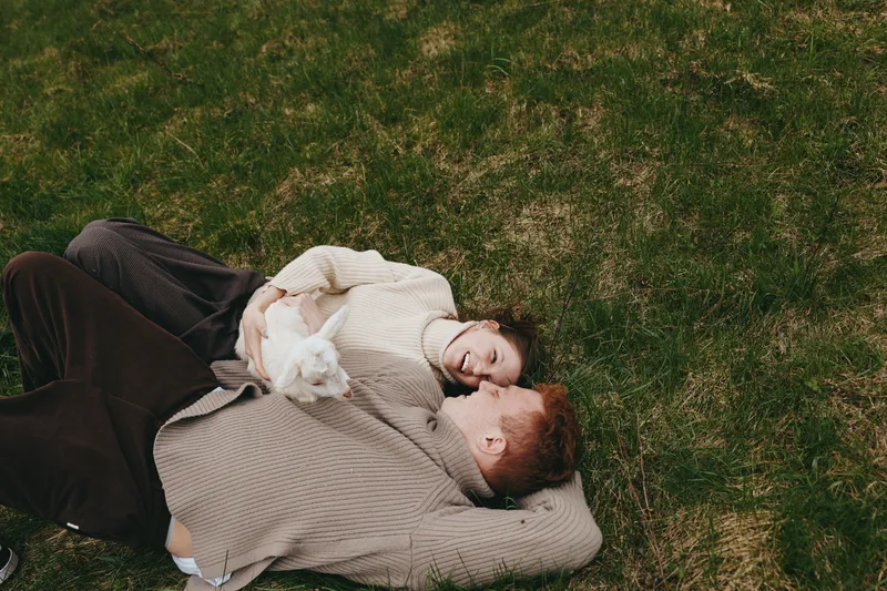 Young couple lying on grass in casual sweaters enjoy a playful moment with a small white baby goat in a relaxed outdoor lifestyle scene during spring.
