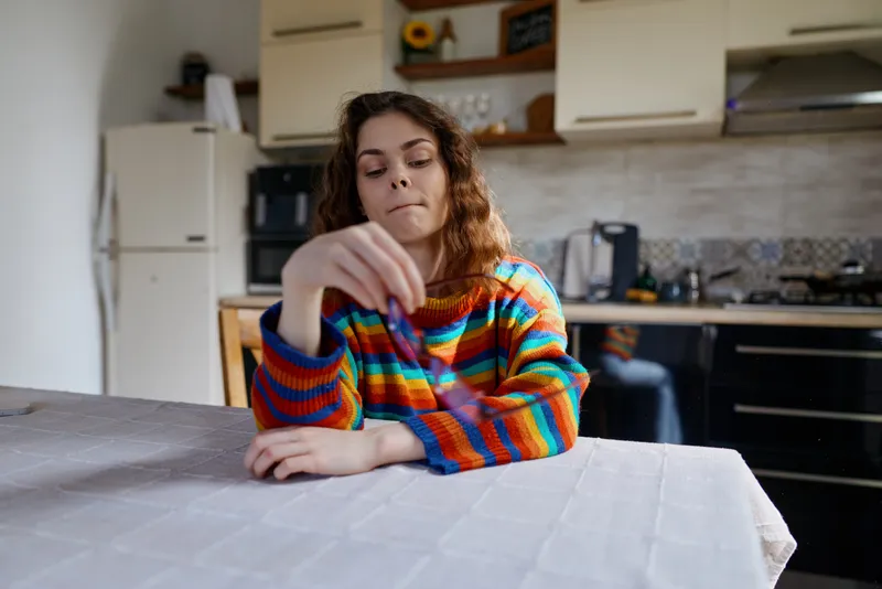 A young woman in a multicolored striped sweater sits at a dining table in a bright kitchen, resting her chin on her hand in a moment of quiet reflection.