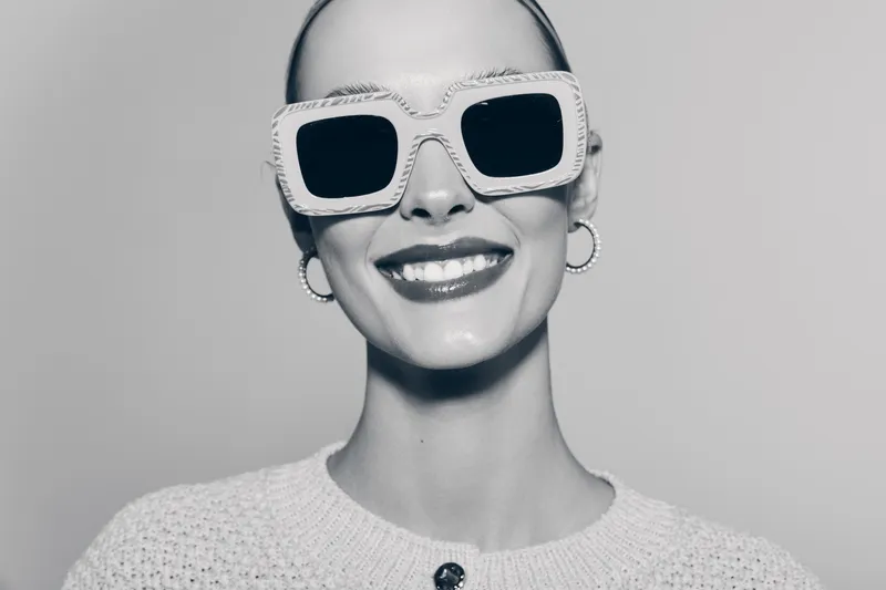 Smiling Caucasian woman with pale skin wearing large white sunglasses and hoop earrings. Close-up portrait in black and white with bright expression on grey background.