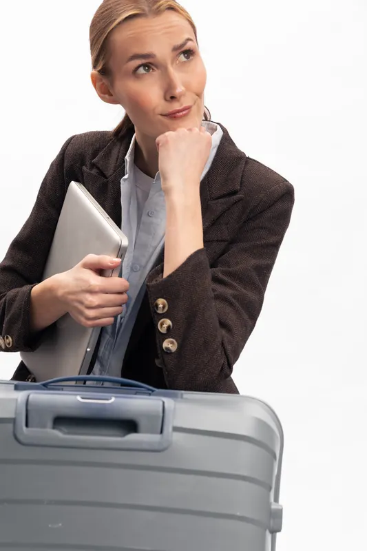 Thoughtful businesswoman in blazer holds laptop and suitcase, looking up with hand on chin. Concept of travel planning, business trip, and professional decision making on white background.