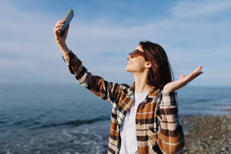 Woman taking a selfie by the sea, wearing a plaid shirt and sunglasses, arms raised in a joyful moment, clear blue sky and calm water behind, outdoors.