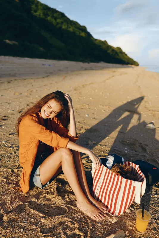 Cheerful woman on a sunlit beach, casual orange shirt, barefoot, relaxed pose, striped bag beside her, soft breeze, warm tones, distant hills, sea in the background