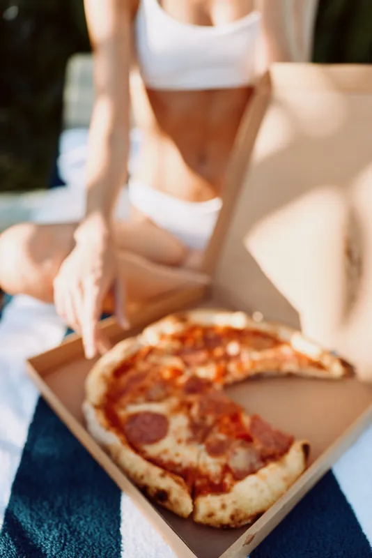 Casual outdoor lifestyle scene shows a person in a white top enjoying pizza from an open box on a blanket, highlighting relaxed moments, shared food, and carefree summer vibes in daylight