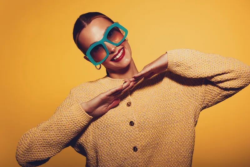 Smiling woman in oversized turquoise sunglasses and beige knitted sweater poses with hands near face on yellow background. Concept of fashion, style, and joyful mood in studio portrait.