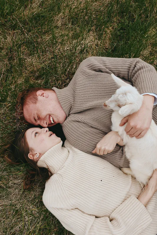 Couple outdoors on grass, lying close and smiling, two cozy sweaters, holding a white cat, sunlit calm moment, relaxed affection, natural colors, intimate lifestyle scene