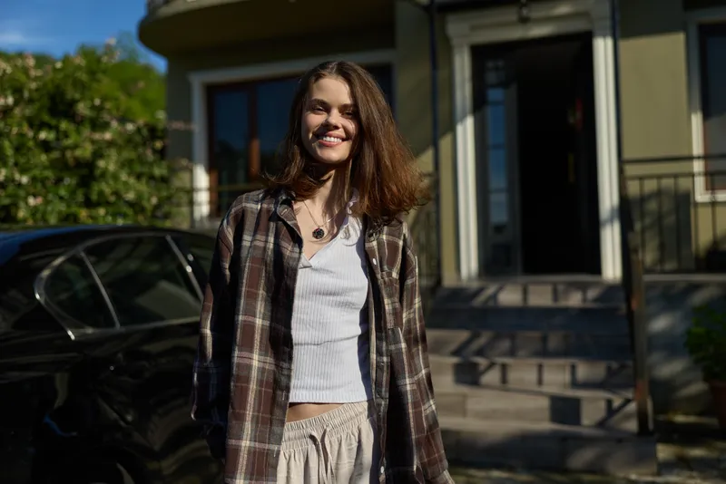 Vibrant woman outdoors, smiling with relaxed posture, casual lifestyle moment in natural light, plaid jacket and jeans, greenery backdrop, sunlit mood, candid feel