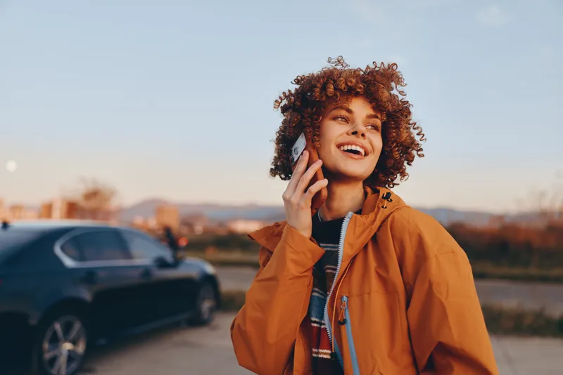 Woman lifestyle portrait outdoors, curly hair orange jacket smiling while speaking on phone, car parked behind, evening sky and suburban road, calm communication concept
