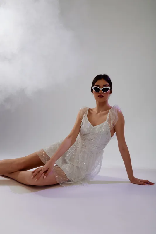 Young woman in a stylish white dress and sunglasses poses on the floor against a neutral background. Concept of beauty, fashion, and modern elegance.