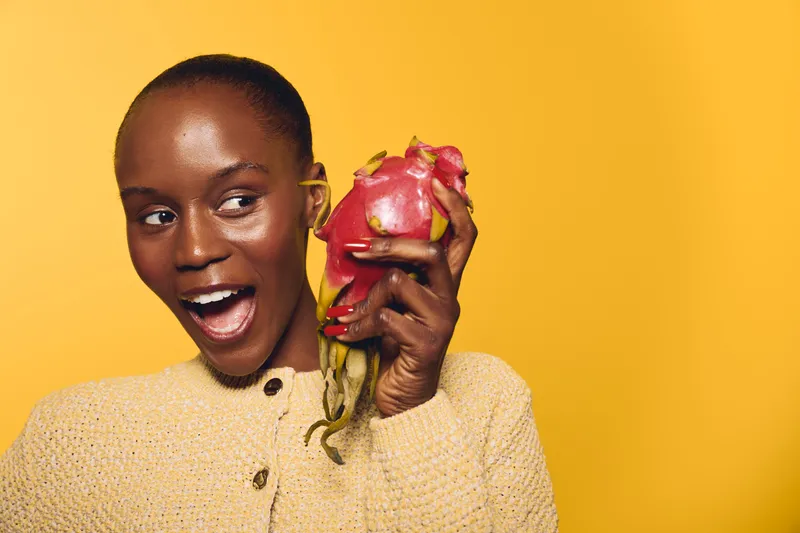 Happy African woman with dark skin holds a ripe dragon fruit near her face, smiling with excitement. Yellow background and cozy beige sweater create a warm, vibrant atmosphere.