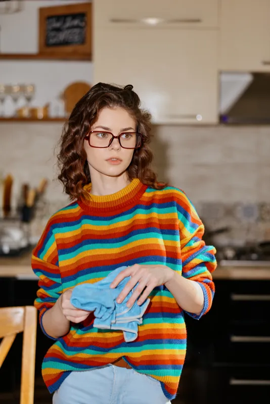 A young woman with curly hair and red glasses stands in a bright kitchen, folding a blue cloth in her hands, conveying a casual, cozy home moment of everyday life, and a sense of personal routine.