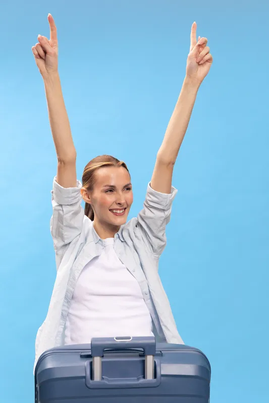 Happy woman with suitcase raised arms up smiling in front of blue background. Concept of travel, vacation, success, joy, and freedom with casual clothes and positive emotions.