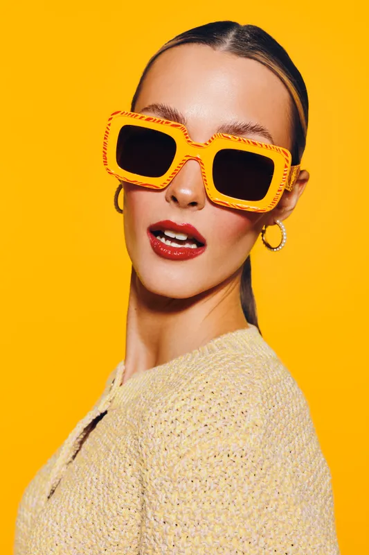 Stylish woman with slicked hair wears large orange sunglasses and yellow sweater against bright yellow background. Fashion portrait with bold colors and confident expression.