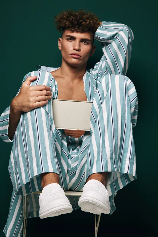 Young man with curly hair wearing striped pajamas and white sneakers sitting on chair holding blank tablet with serious expression on dark green background.