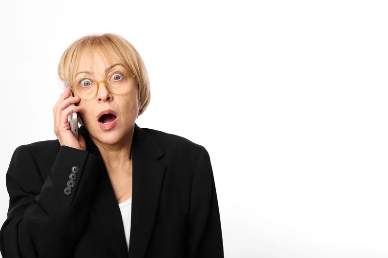 Woman in business attire holding smartphone near ear, phone call, shocked surprised face, glasses visible, office communication studio portrait on white background, copy space