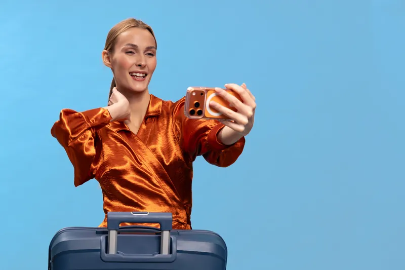 Young woman in shiny orange blouse takes selfie with smartphone while standing behind blue suitcase on light blue background. Concept for travel, vacation, tourism, and lifestyle.