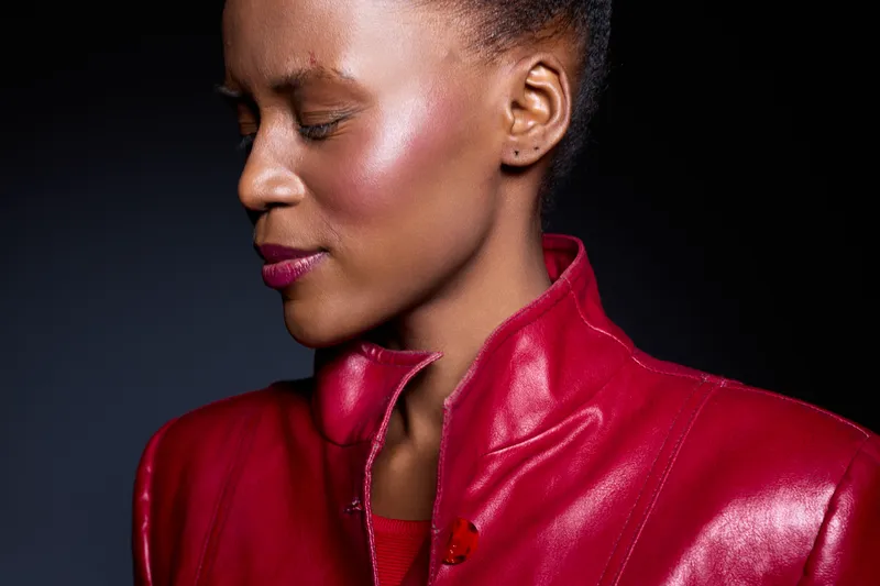 Close-up of an elegant dark skinned African woman with closed eyes wearing a red leather jacket against black background. Concept of fashion, beauty, and confidence.