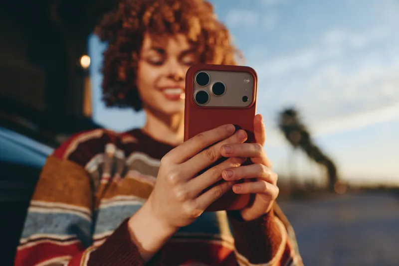 Woman lifestyle selfie with smartphone, curlyhair woman smiling while holding phone, warm golden sunset on city street background, striped sweater, evening outdoors, shallow depth of field