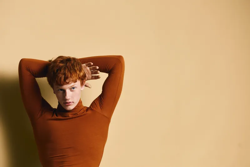 Redhead woman posing with hands behind head wearing brown turtleneck sweater against plain beige background in studio for fashion portrait photo.