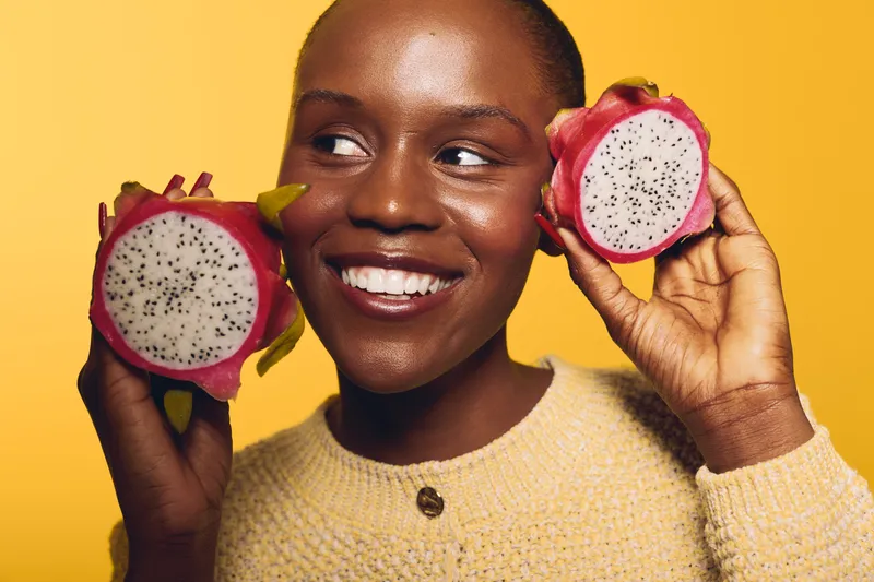 Smiling black woman holds two halves of dragon fruit near her face on yellow background. Concept of healthy lifestyle, fresh fruit, natural food, and positive emotions with dark skin model.